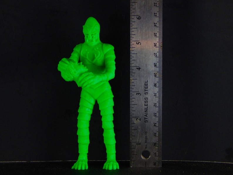 Pu&ograve; includere: Una action figure di plastica verde di una creatura con un corpo squamoso e testurizzato e una grande testa. La figura tiene una figura verde pi&ugrave; piccola nelle sue mani.