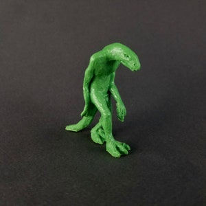 May include: Green 3D printed figurine of a humanoid creature with reptilian features. The creature has a long tail, long arms, and a long, thin neck. It is standing on two legs and has a slightly hunched posture.