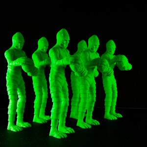 Pu&ograve; includere: Cinque figure di plastica verde di una creatura di un film horror. Le figure sono in piedi in fila, rivolte in avanti. Hanno tutte la stessa posa, con le braccia alzate e le mani chiuse a pugno.