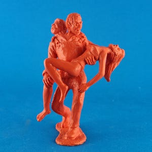 May include: A monochromatic orange figurine depicting a three-headed figure carrying a woman. The figures are stylized with exaggerated features, set against a solid blue background. The figurine appears to be made of a solid material.