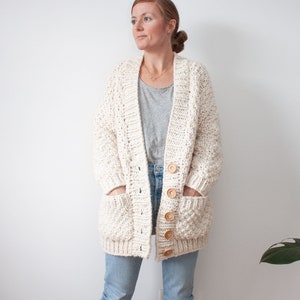 Peut inclure: Un cardigan en crochet blanc avec des boutons en bois et des poches. Le cardigan est porté sur un t-shirt gris et un jean bleu.