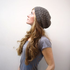 Puede incluir: Una mujer con el pelo largo y ondulado castaño lleva una boina de punto gris. Está mirando hacia arriba y hacia la derecha.