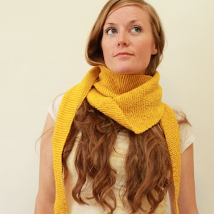 May include: A mustard yellow knitted scarf is draped around the neck. The scarf is made of a thick yarn and has a triangular shape. The ends of the scarf hang down. The person in the image is wearing a white t-shirt.
