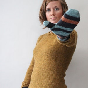 Può includere: Una donna indossa un maglione giallo senape con un paio di guanti senza dita a righe blu e marroni.