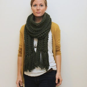 Op de afbeelding: Een vrouw draagt een mosterdgele cardigan over een wit t-shirt en een donkerblauwe jeans. Ze draagt een donkergroene gehaakte sjaal met lange franjes.