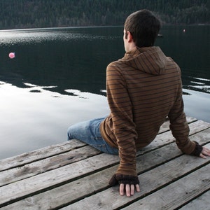 Könnte beinhalten: Eine Person in einem braunen und schwarzen gestreiften Kapuzenpullover sitzt auf einem Holzsteg und blickt auf einen See. Das Wasser ist ruhig und spiegelt die umliegenden Bäume wider.