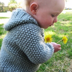Op de afbeelding: Een jong kind draagt een lichtblauwe gebreide trui met capuchon met een textuurpatroon en houdt twee gele paardenbloemen vast.