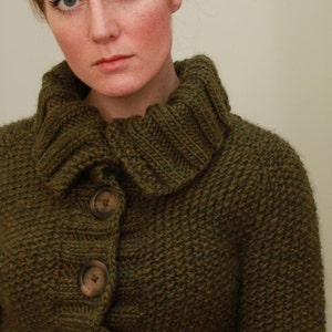Può includere: Primo piano di una donna che indossa un cardigan di maglia verde scuro con collo alto e chiusura con bottoni.