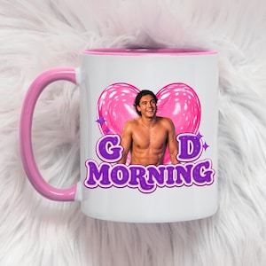 May include: A white ceramic mug with a pink handle and interior rim. The front features a graphic of a shirtless person in front of a large pink heart, with the purple text "GOOD MORNING" written below, accented by small purple star icons.