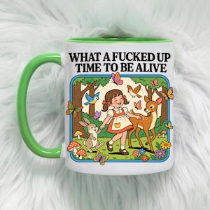 Può includere: Una tazza in ceramica bianca con manico e bordo verde brillante, caratterizzata da un'illustrazione colorata di un bambino in un bosco con un cervo, un coniglio, uccelli e farfalle. Il testo "WHAT A FUCKED UP TIME TO BE ALIVE" è stampato in nero.