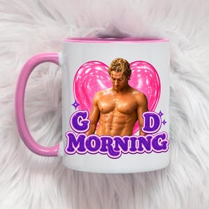 May include: A white ceramic mug with a pink handle and interior rim. The front features a graphic of a shirtless man in front of a large pink heart, with the purple text "G D MORNING" printed below the image, accented by small purple star icons.
