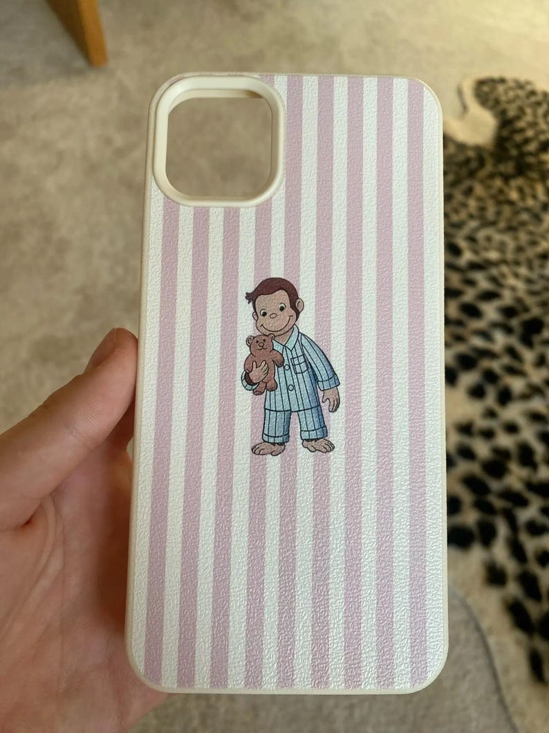 May include: White phone case with vertical pink stripes. The case features a cartoon illustration of a monkey in blue pajamas holding a teddy bear. The case has a camera cutout and a white border.