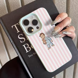 May include: A phone case featuring a cartoon illustration of a boy in pajamas holding a teddy bear. The case has a pink and white striped pattern and a light blue camera frame. The case is resting on a black book.