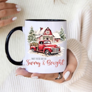 Könnte beinhalten: Ein weißer Keramikbecher mit schwarzem Henkel und Rand, verziert mit einer Aquarellillustration eines roten Pick-ups vor einer verschneiten Scheune und Tannenbäumen. Der Text lautet May Your Day Be Farmy & Bright in roter Schrift.