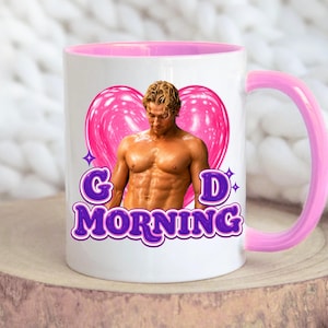May include: A white ceramic mug with a pink interior and handle. The front features a graphic of a shirtless person with blonde hair in front of a large pink heart. Below the image, the purple text reads G D MORNING with small star accents.