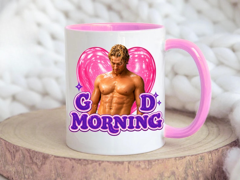 May include: A white ceramic mug with a pink interior and handle. The front features a graphic of a shirtless person with blonde hair in front of a large pink heart. Below the image, the purple text reads G D MORNING with small star accents.