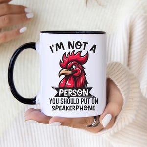 Peut inclure: Un mug en céramique blanche avec une anse et un bord noirs, orné d'un dessin de tête de coq rouge au-dessus du texte noir "I'M NOT A PERSON YOU SHOULD PUT ON SPEAKERPHONE" imprimé en caractères gras sans empattement sur la face avant.