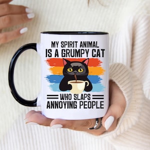 Op de afbeelding: Een witte keramische mok met een zwarte handgreep en rand, voorzien van een afbeelding van een zwarte kat die een klein kopje vasthoudt. De tekst luidt: MY SPIRIT ANIMAL IS A GRUMPY CAT WHO SLAPS ANNOYING PEOPLE. Het ontwerp bevat horizontale rode, oranje en blauwe strepen.