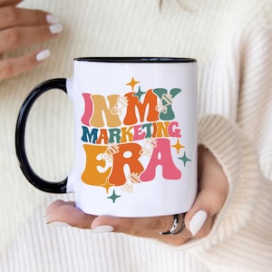 Op de afbeelding: Een witte keramische mok met een zwarte handgreep en rand, voorzien van de kleurrijke tekst "IN MY MARKETING ERA" in een retro, golvend lettertype. Het ontwerp bevat kleine megafoonpictogrammen en vierpuntige sterren in oranje, roze, blauwgroen en geel.