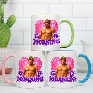 May include: Three white ceramic mugs with colored handles and interiors. Each mug features a graphic of a shirtless person in front of a pink heart, with the purple text "GD MORNING" below. The handles are light green, light pink, and light blue.