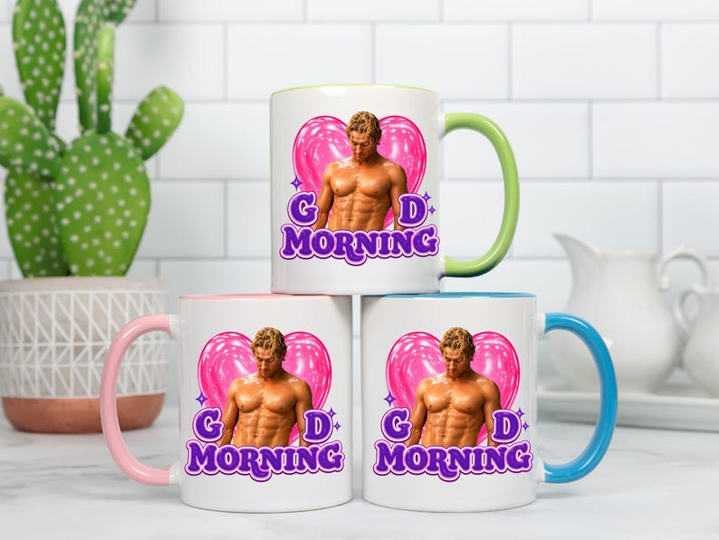 May include: Three white ceramic mugs with colored handles and interiors. Each mug features a graphic of a shirtless person in front of a pink heart, with the purple text "GD MORNING" below. The handles are light green, light pink, and light blue.