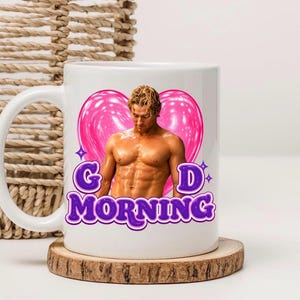 May include: A white ceramic mug featuring a graphic of a shirtless person with blonde hair positioned in front of a large, glossy pink heart. Below the figure, the text "G D MORNING" is printed in bold, purple, bubble-style lettering with white outlines.