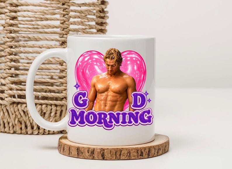 May include: A white ceramic mug featuring a graphic of a shirtless person with blonde hair positioned in front of a large, glossy pink heart. Below the figure, the text "G D MORNING" is printed in bold, purple, bubble-style lettering with white outlines.