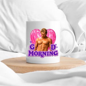 May include: A white ceramic mug featuring a graphic of a shirtless person with blonde hair positioned in front of a large pink heart. The text "G D MORNING" is printed in bold purple letters below the heart, accented by small purple star icons.