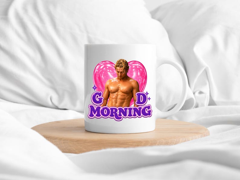 May include: A white ceramic mug featuring a graphic of a shirtless person with blonde hair positioned in front of a large pink heart. The text "G D MORNING" is printed in bold purple letters below the heart, accented by small purple star icons.
