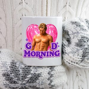 May include: A white ceramic mug featuring a graphic of a shirtless person with a muscular build positioned in front of a large pink heart. The text G D MORNING is printed in purple letters with a white outline below the heart and the person.