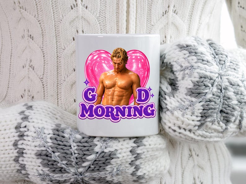 May include: A white ceramic mug featuring a graphic of a shirtless person with a muscular build positioned in front of a large pink heart. The text G D MORNING is printed in purple letters with a white outline below the heart and the person.