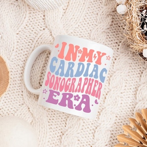 Könnte beinhalten: Ein weißer Keramikbecher mit dem Schriftzug IN MY CARDIAC SONOGRAPHER ERA in einer wellenförmigen Retro-Schriftart. Die Beschriftung verwendet eine Farbpalette aus Rosa, Hellblau und Lila, akzentuiert durch kleine grafische Elemente.