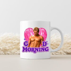 May include: A white ceramic mug featuring a graphic of a shirtless person with blonde hair positioned in front of a large, glossy pink heart. The text "GD MORNING" is printed in bold, purple, outlined letters below the heart and figure.
