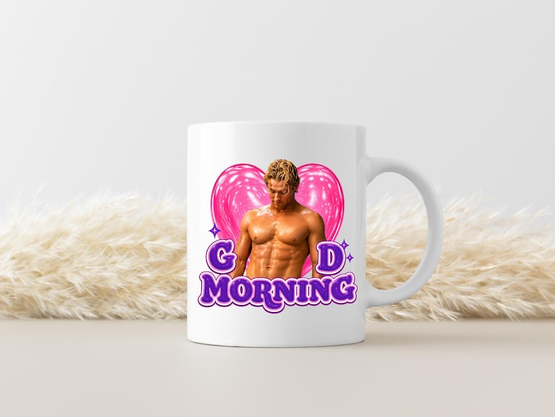 May include: A white ceramic mug featuring a graphic of a shirtless person with blonde hair positioned in front of a large, glossy pink heart. The text "GD MORNING" is printed in bold, purple, outlined letters below the heart and figure.