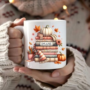 Può includere: Una tazza in ceramica bianca con un'illustrazione colorata di una pila di libri, una zucca bianca, una zucca arancione, foglie autunnali e un portamatite. Il testo "Just a girl who loves fall" è stampato sul dorso di uno dei libri.