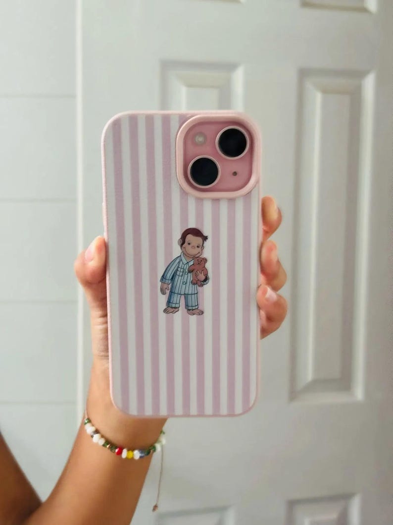 May include: Pink and white striped phone case with a cartoon illustration of a character in pajamas holding a teddy bear. The case has a pink camera module and a matching pink border.