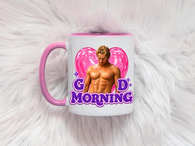 May include: A white ceramic mug with a pink handle and interior rim. The front features a graphic of a shirtless man in front of a large pink heart, with the purple text "G D MORNING" printed below the image, accented by small purple star icons.