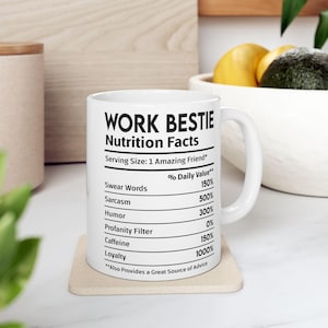 Op de afbeelding: Een witte keramische mok met zwarte tekst die Work Bestie Nutrition Facts leest, met verschillende percentages voor scheldwoorden, sarcasme, humor, profaniteitsfilter, cafeïne en loyaliteit, met een opmerking over het geven van advies.