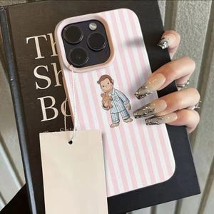 May include: Pink and white striped phone case featuring a cartoon character in pajamas holding a teddy bear. The case has a light pink border and a camera cutout. A blank white tag hangs from the case.