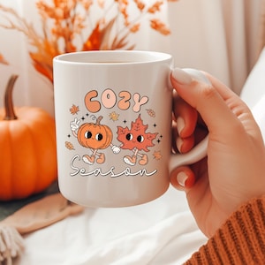 May include: A white ceramic mug featuring a graphic illustration of a cartoon pumpkin and a maple leaf holding hands. The text "COZY" appears in orange above the characters, and "Season" is written in black cursive script below them.
