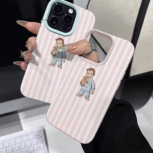 May include: Two phone cases with a pink and white striped pattern. Each case features a cartoon illustration of a boy in pajamas holding a teddy bear. The cases have a light blue and pink border.
