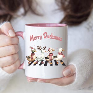 Könnte beinhalten: Ein weißer Keramikbecher mit rosafarbenem Innenraum und Henkel, bedruckt mit vier Enten, die über einen schwarz-weiß gestreiften Zebrastreifen laufen. Die Enten tragen winterliche Accessoires und der Schriftzug Merry Ducksmas ist in Rosa.