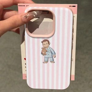 May include: Pink and white striped phone case featuring a cartoon illustration of a monkey in pajamas holding a teddy bear. The case has a pink border and is shown with a pink paper backing.