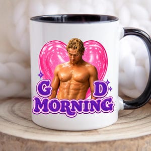 May include: A white ceramic mug with a black interior and handle. The front features a graphic of a shirtless man with blonde hair centered in a pink heart, with the purple text "G D MORNING" printed below the heart and the man's torso.