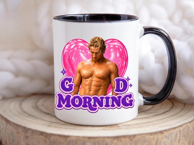 May include: A white ceramic mug with a black interior and handle. The front features a graphic of a shirtless man with blonde hair centered in a pink heart, with the purple text "G D MORNING" printed below the heart and the man's torso.