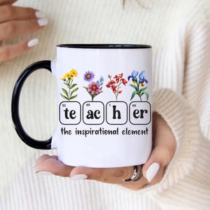 Op de afbeelding: Een witte keramische mok met een zwarte handgreep en rand. Het ontwerp toont vier vierkanten in de stijl van het periodiek systeem met de letters 'te', 'ac', 'h' en 'er', bekroond met kleurrijke bloemenillustraties. Daaronder staat de tekst 'the inspirational element'.