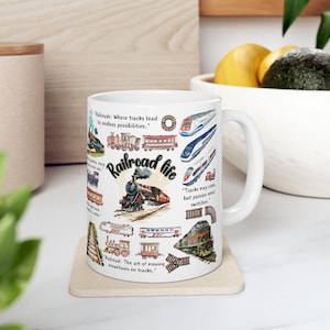 Puede incluir: Una taza de cerámica blanca con un collage colorido de varios trenes, incluyendo locomotoras de vapor y modernas, junto con el texto Railroad life y varias citas inspiradoras sobre trenes y vías impresas en letras negras.