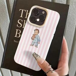 May include: A pink and white striped phone case featuring a cartoon illustration of a monkey holding a teddy bear. The phone case has a white border and a camera cutout. The case is resting on a dark book.
