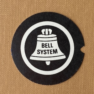 May include: Black and white round coaster with a white bell and the text "BELL SYSTEM" inside a white circle.