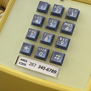 May include: Close-up of a vintage yellow telephone keypad with gray number and letter keys. The keypad includes numbers 1-9, the asterisk, the pound sign, and the word "OPER" for the number 0. The area code 267 and phone number 345-6789 are also visible.